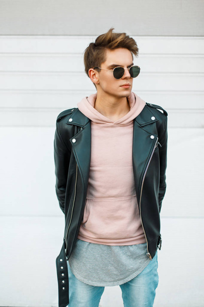 Komea nuori muodikas mies, jolla on hiusten leikkaus aurinkolaseissa nahkaisessa mustassa takissa ja vaaleanpunainen huppari lähellä valkoista metalliseinää.
 - Valokuva, kuva