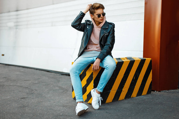 Handsome young model of a man wearing sunglasses in a black leather jacket, a pink sweatshirt, blue jeans and white shoes sits on a black-and-yellow slab - Foto, Imagen