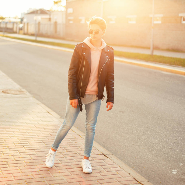 Jovem elegante em óculos de sol vintage em uma jaqueta de couro preto, jeans azuis e tênis brancos posando ao pôr do sol
 - Foto, Imagem