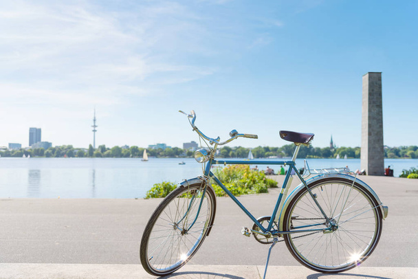 bicicleta azul vintage estacionada en la orilla del lago Alster en Hamburgo, Alemania bajo un hermoso cielo azul claro de verano con paisaje urbano en un fondo borroso
 - Foto, Imagen