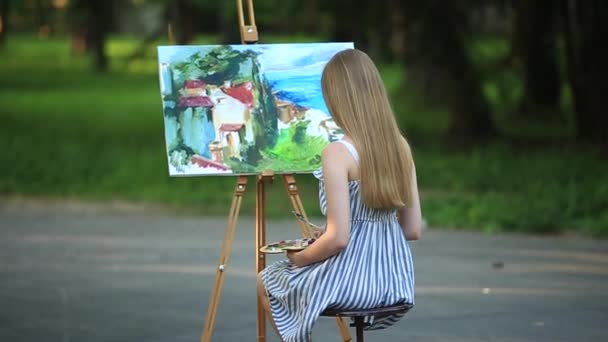 Hermosa chica dibuja un cuadro en el parque usando una paleta con pinturas y una espátula. caballete y lienzo con una imagen
. - Metraje, vídeo