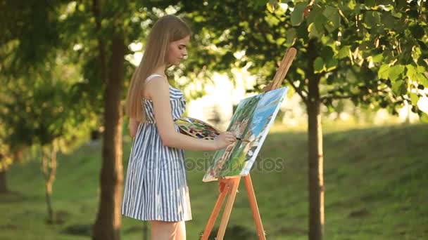 Όμορφο κορίτσι τραβάει μια εικόνα στο πάρκο χρησιμοποιώντας μια παλέτα με χρώματα και μια σπάτουλα. Καβαλέτο και καμβάς με την εικόνα. - Πλάνα, βίντεο