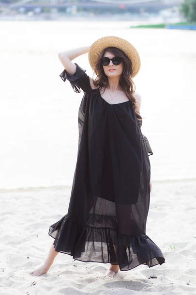 Красивая юная брюнетка в черном платье, черные очки и соломенная шляпа, гуляющая по песку на пляже и позирующая на рассвете летом
 - Фото, изображение