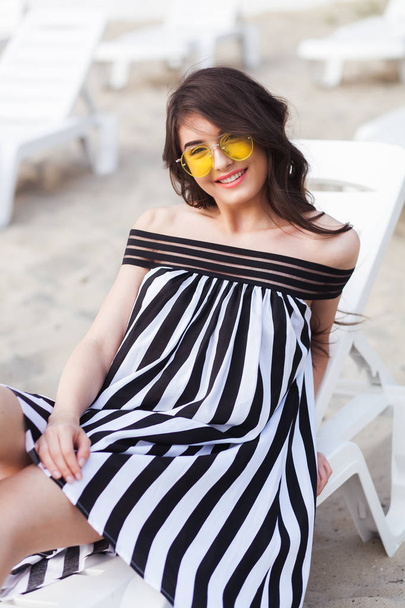 サンベッドのビーチでリラックスしながら座っている黒と白のストライプのドレスで幸せな若いブルネットの女性の肖像画 - 写真・画像