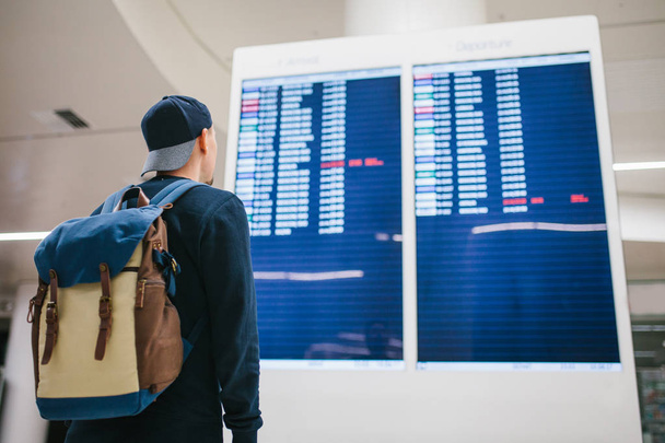 Een jonge mannelijke reiziger met een rugzak in casual stijl wordt gekeken naar het informatiebord op de luchthaven. Informatie opvragen over de vlucht. Overdracht. - Foto, afbeelding