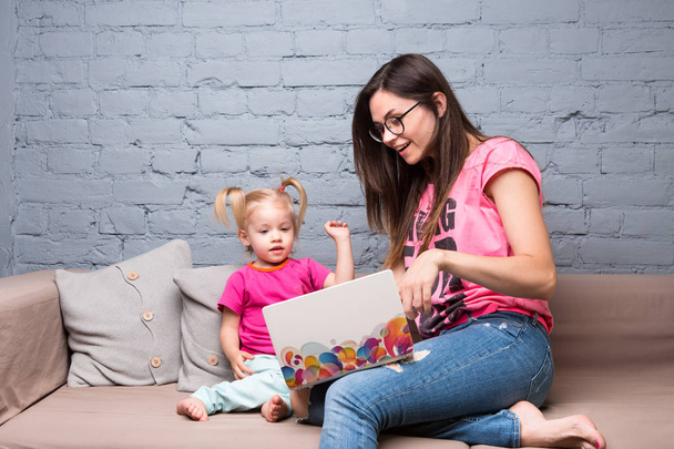 Joven madre y niña de dos años con el pelo rubio sentado en el sofá y el uso de la computadora portátil. Vestido con ropa rosa brillante
 - Foto, imagen