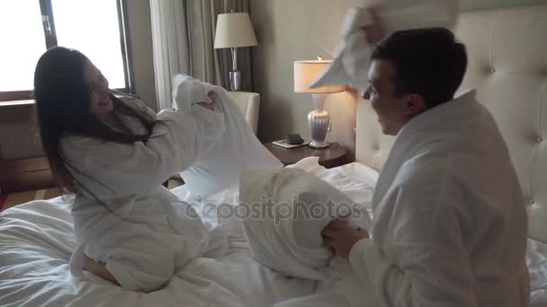 Milující pár v bílých pláštích na postel šťastně bojuje s polštáři zpomalené stopáže videa - Záběry, video