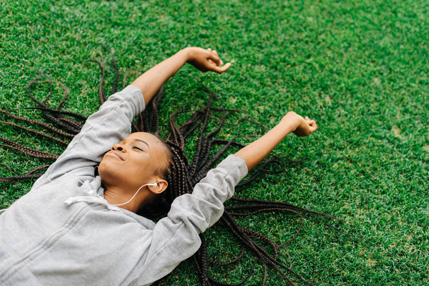 Delighted afro-american teenager is enjoying music while laying on the grass and raising hands. - Photo, Image
