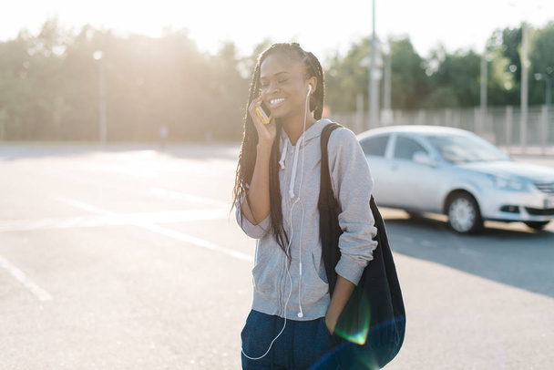 Half-length portrait of the cheerful afro-american teenager talking via the mobile phone while walking along the street during the sunset. - 写真・画像