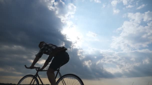 Σιλουέτα του νεαρού άνδρα ιππασία στο ρετρό ποδήλατο με όμορφο ηλιοβασίλεμα ουρανό στο υπόβαθρο. Αθλητικός τύπος ποδηλασία στον δρόμο χώρας. Αρσενικό ποδηλάτης ιππασίας ποδήλατο. Δραστήριου τρόπου ζωής αργή κίνηση - Πλάνα, βίντεο