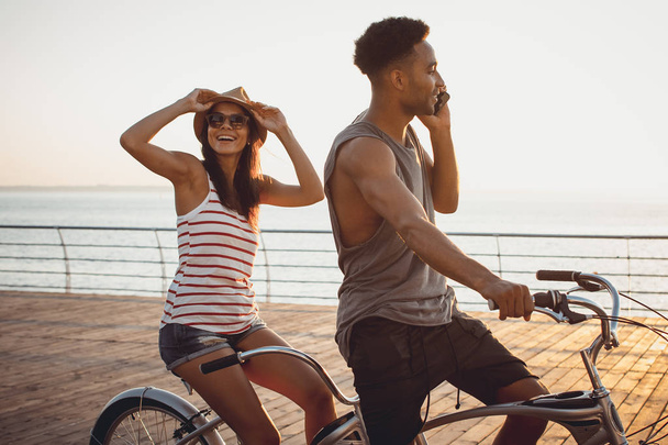 Portrait of a mixed race couple on tandem bicycle outdoors near the sea, young african black man talking on phone - Foto, Imagen