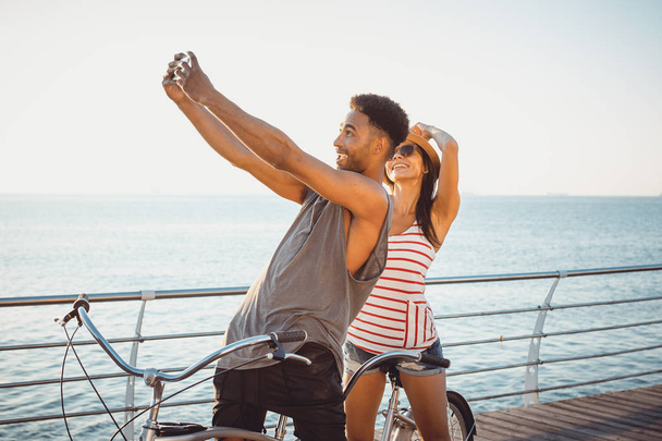 Portrait of a mixed race couple doing selfie on tandem bicycle outdoors near the sea - Фото, зображення