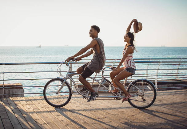 Portrait of a mixed race couple on tandem bicycle outdoors near the sea - Fotó, kép