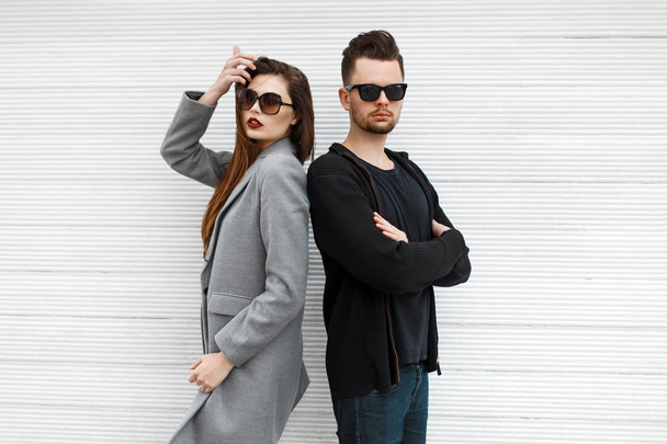 Hermosa pareja de moda con gafas de sol en ropa moderna con estilo posando cerca de una pared de madera blanca
. - Foto, imagen