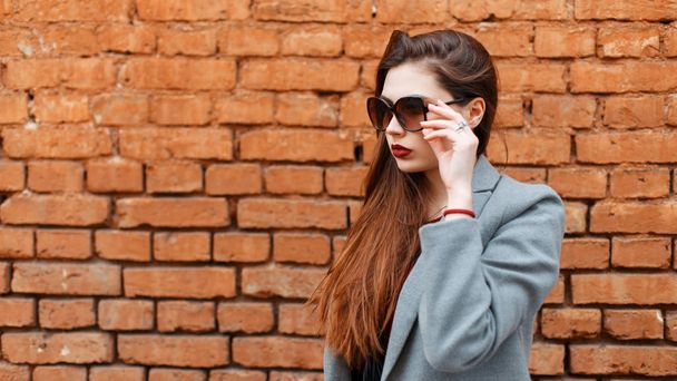 Elegante joven hermosa mujer con gafas de sol en un abrigo gris cerca de una pared de ladrillo
 - Foto, Imagen
