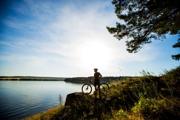 岩の上に立っていると、夕日を楽しむ自転車ライダーの側面図です。動機と冒険のためのインスピレーション. - 写真・画像