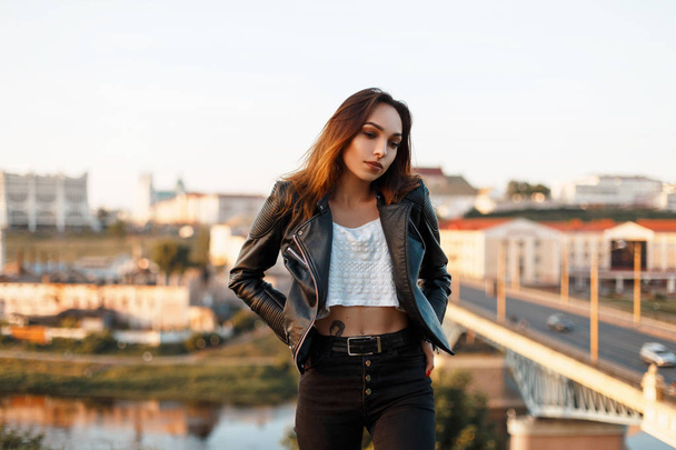 Beautiful young woman in a fashionable leather black jacket and white tank top, jeans vintage jeans urban background - Photo, Image