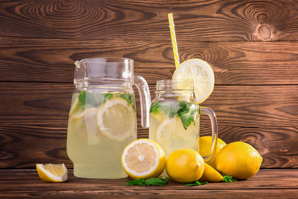 A huge pitcher and a mason jug full of refreshing citrus lemonade or mojito. Glasses of summer cocktails with ripe and juicy lemons and fresh green mint on a dark brown wooden table.  - Foto, Imagen