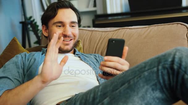 Giovane uomo attraente che chatta con gli amici in skype utilizzando smartphone mentre si rilassa sul divano a casa
. - Filmati, video