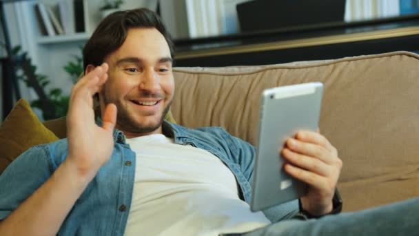 Портрет щасливого привабливого чоловіка, який спілкується з сім'єю в скайпі, використовуючи планшет, лежачи на дивані вдома
. - Кадри, відео