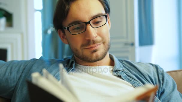 Jovem homem atraente com óculos relaxantes no sofá em casa e livro de leitura. Fechar tiro
. - Filmagem, Vídeo