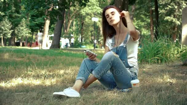 brunette listens to music with headphones on the phone sitting on the grass - Filmati, video