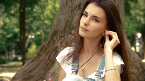 beautiful young lady with headphones in your ears, sitting near a tree and listens to music - Filmati, video