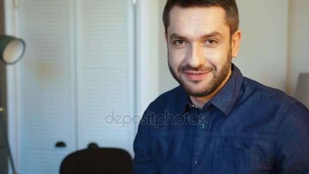 Portrait of caucasian handsome man with beard in blue shirt putting glasses, looking to the camera and smiling. Indoor. - Footage, Video