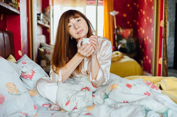 Porträt einer Frau mittleren Alters mit roten Haaren, die am sonnigen Morgen auf dem Bett sitzt und eine weiße Tasse Tee in der Hand hält. - Foto, Bild