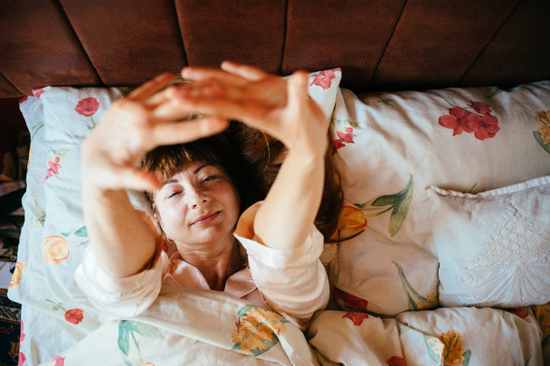 Вид зверху чарівна жінка середнього віку з червоно-коричневим волоссям у сплячому одязі повільно прокидається в ліжку
. - Фото, зображення