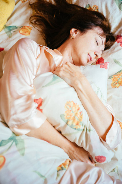 Bovenaanzicht van mooie midden leeftijd vrouw met rood haar slapen en knuffelen het kussen op het bed in zonnige ochtend. - Foto, afbeelding