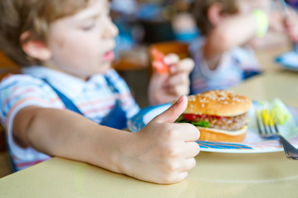Lindo niño preescolar saludable come hamburguesa sentada en el comedor de la escuela
 - Foto, imagen