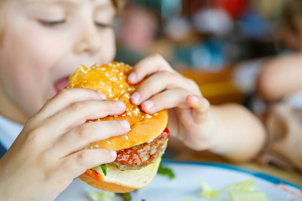 Ładny chłopak przedszkola zjada hamburger siedzi w stołówce szkolnej - Zdjęcie, obraz