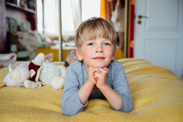 Милый маленький блондин лежит на животе на кровати и смотрит вперед, большой голубые глаза мальчика интересно
. - Фото, изображение