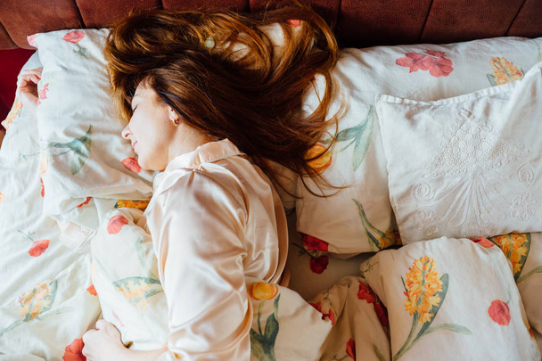 Vista superior de la encantadora mujer de mediana edad con el pelo marrón rojo en ropa de dormir rosa durmiendo lentamente en la cama
. - Foto, imagen