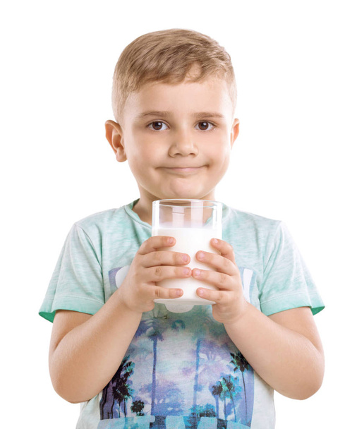 A handsome little boy holding a glass of milk in his hands isolated on a white background. The beautiful boy is wearing a blue T-shirt and is standing with a drain of fresh milk. - Photo, Image