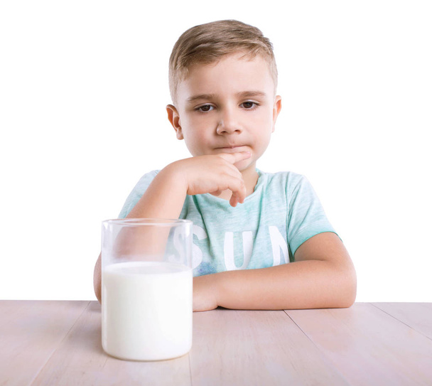 A thoughtful and nice little boy in a blue T-shirt with a big glass full of healthy and tasty milk on a wooden table, isolated on a white background. - Fotó, kép