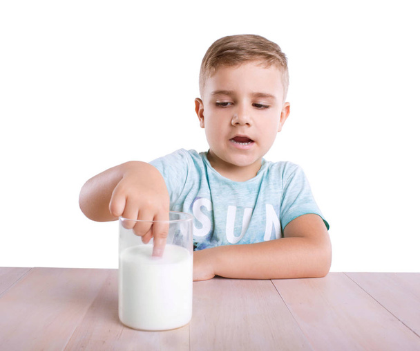 A close-up of a cute little boy is touching a white and organic milk in a transparent glass, which is standing on a wooden table, isolated on a white background.  - Foto, Imagen