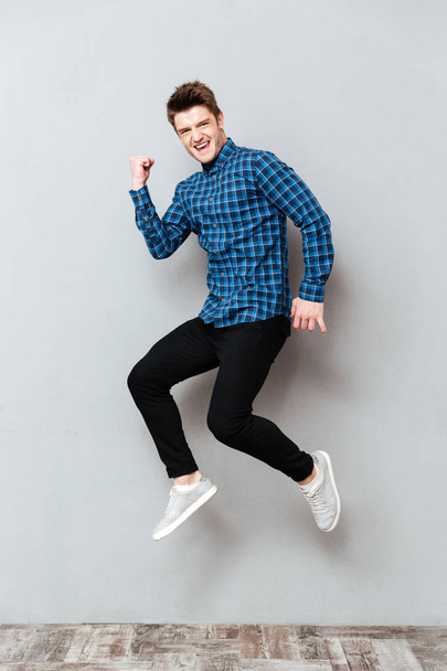 Screaming young man standing over grey wall and jumping. - Фото, зображення