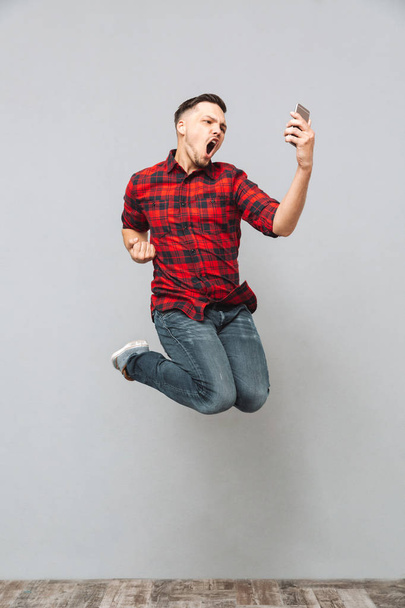 Screaming young man using mobile phone make winner gesture. - Фото, изображение