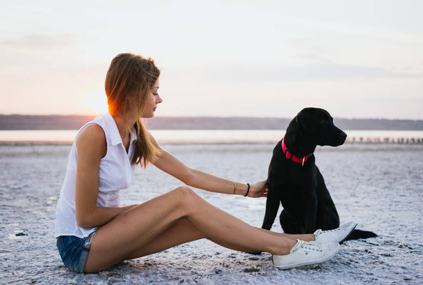 Onnellinen nuori nainen labradorinnoutajan koiran kanssa rannalla auringonlaskun aikaan
 - Valokuva, kuva