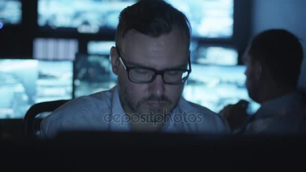 Focused security officer is working on a computer in a dark monitoring room filled with display screens. - Felvétel, videó