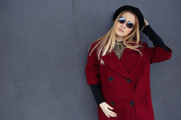 Stylish blonde with beautiful makeup in a red coat, hat and black glasses on the background of a gray wall - Fotó, kép