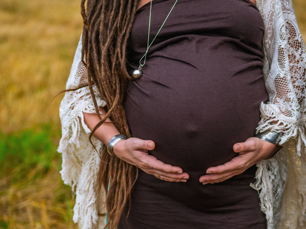 Tummy of pregnant woman in a brown dress with dreadlocks on background of yellow grass field, nature, autumn or summer. Boho style lady - Photo, Image