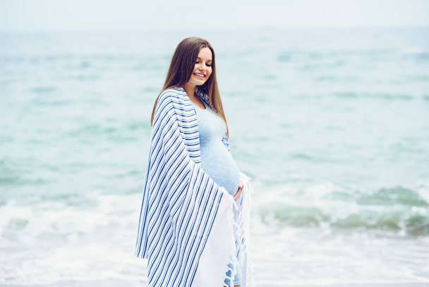 A beautiful pregnant woman in a blue tight dress is covered with a white striped plaid on the beach touching her belly with love and care. Walking on the seashore in sunny day. - Photo, Image
