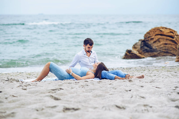 Beautiful pregnant woman and her respectable man leaning against each other and sitting on the seashore. Lovely young couple. - Фото, изображение