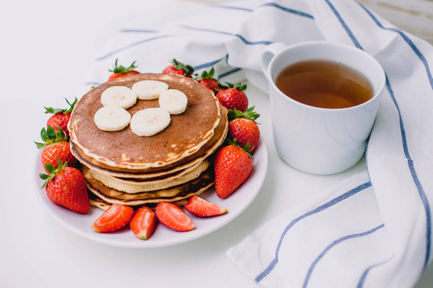 Healthy breakfast. Pancakes with strawberries, bananas, cup of black tea on white background with white towel - Foto, afbeelding