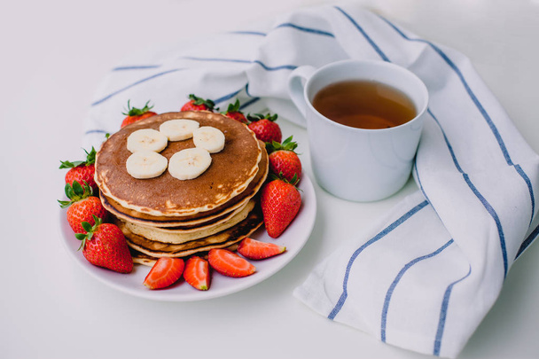 Healthy breakfast. Pancakes with strawberries, bananas, cup of black tea on white background with white towel - Photo, Image
