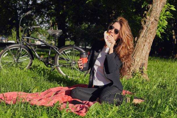 modische junge Frau mit Spiegelglasbrille, formalen Anzug genießen leckeres Gebäck zum Frühstück sitzt an einer roten Decke im Park.hungrige Kaukasier in trendiger schwarzer Jacke stillen Hunger draußen. - Foto, Bild