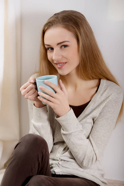 schöne kaukasische Frau trinkt heißen Kaffee oder Tee und schaut durch das Fenster. - Foto, Bild